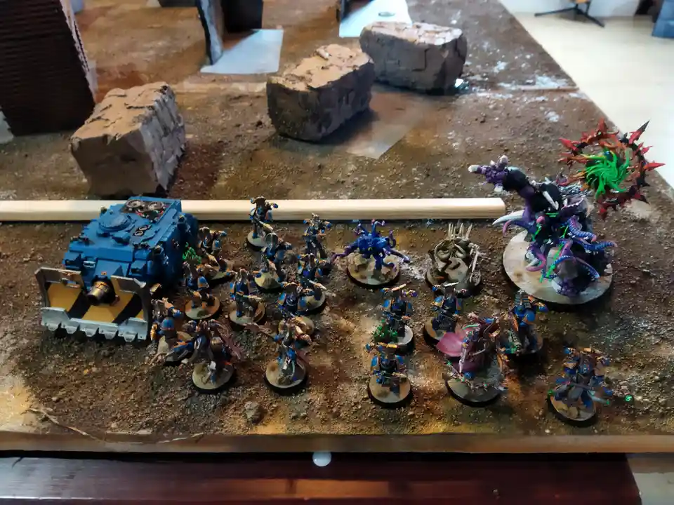 A tabletop wargaming scene shows a large force of blue-painted miniature soldiers and a tank facing off against a single…