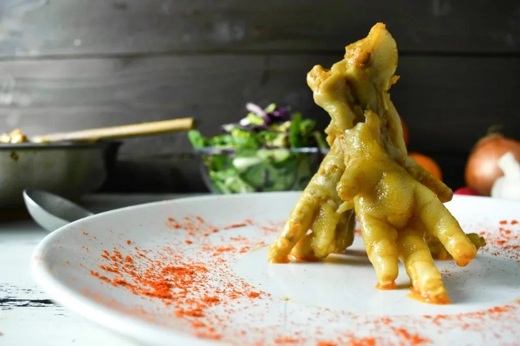 A white plate holds several cooked chicken feet arranged in a tower, dusted with red spice. In the background, a salad bowl…