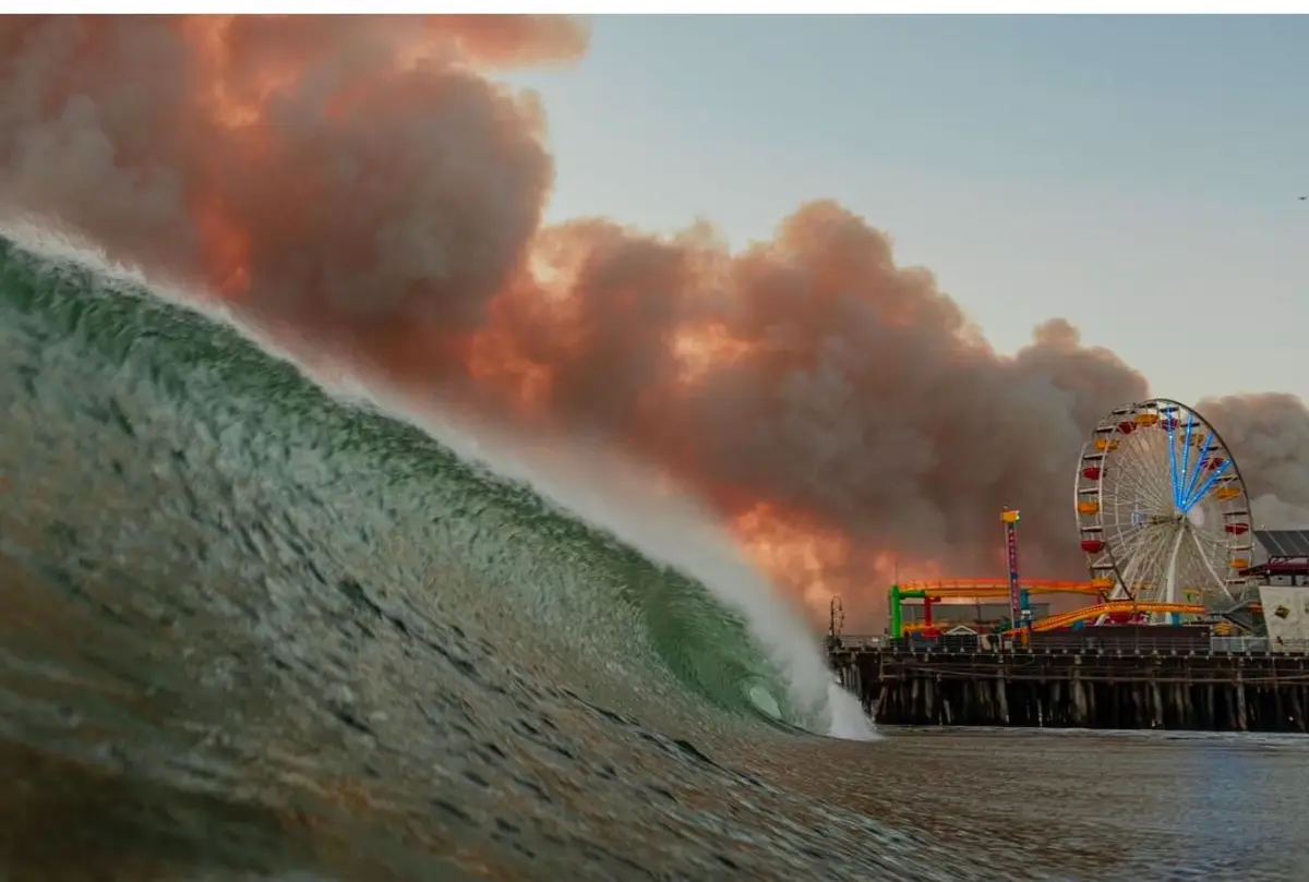 A massive, curling ocean wave dominates the foreground, appearing to crash toward a pier with a Ferris wheel and carnival…