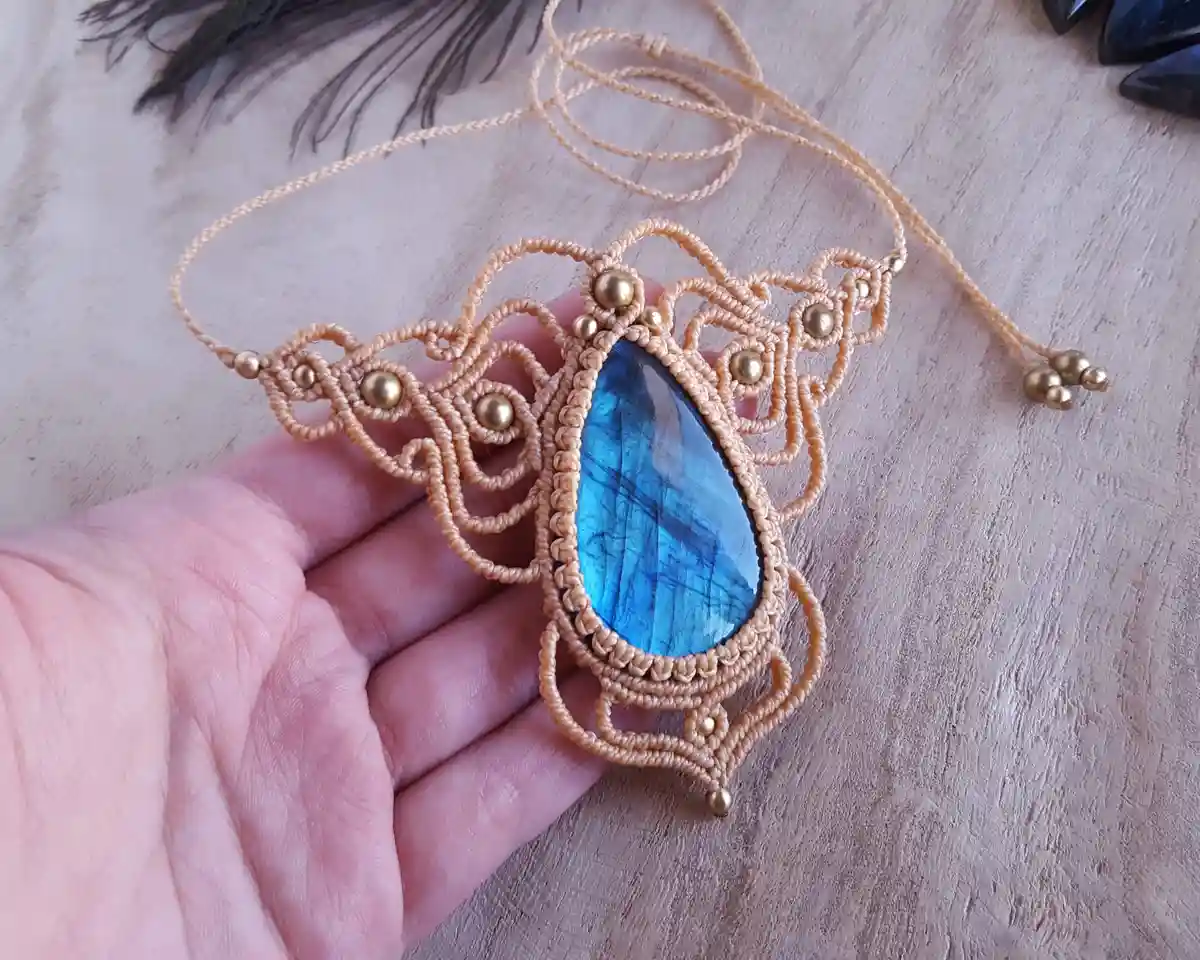 A hand holds an ornate macrame pendant featuring a large, vibrant blue labradorite stone set in a beige cord design with…