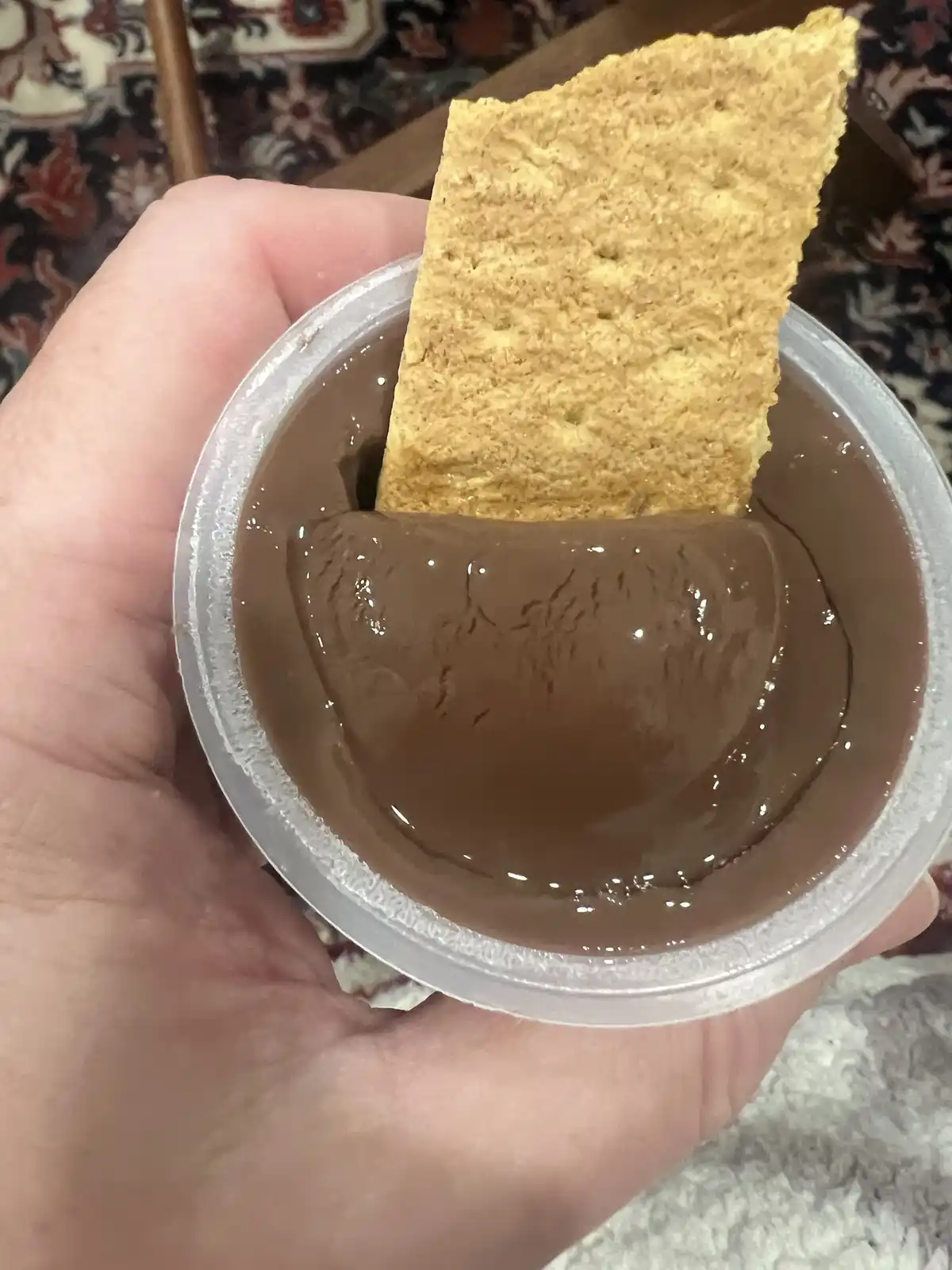 A hand holds a clear plastic cup filled with chocolate pudding, with a rectangular graham cracker partially submerged in it…