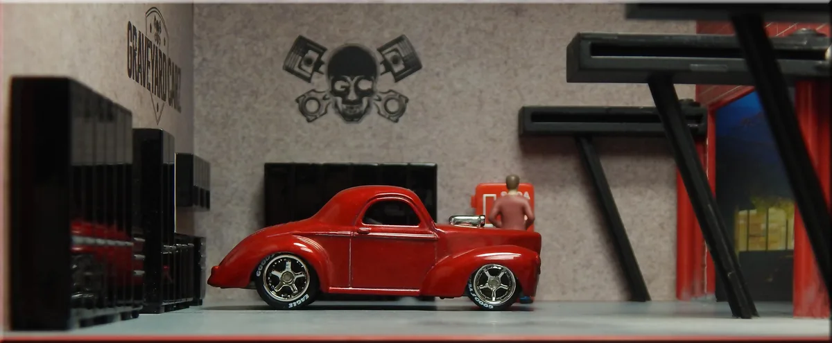 A miniature red hot rod is parked inside a diorama garage labeled 'GRAVEYARD CAR' with a skull and piston logo on the wall…