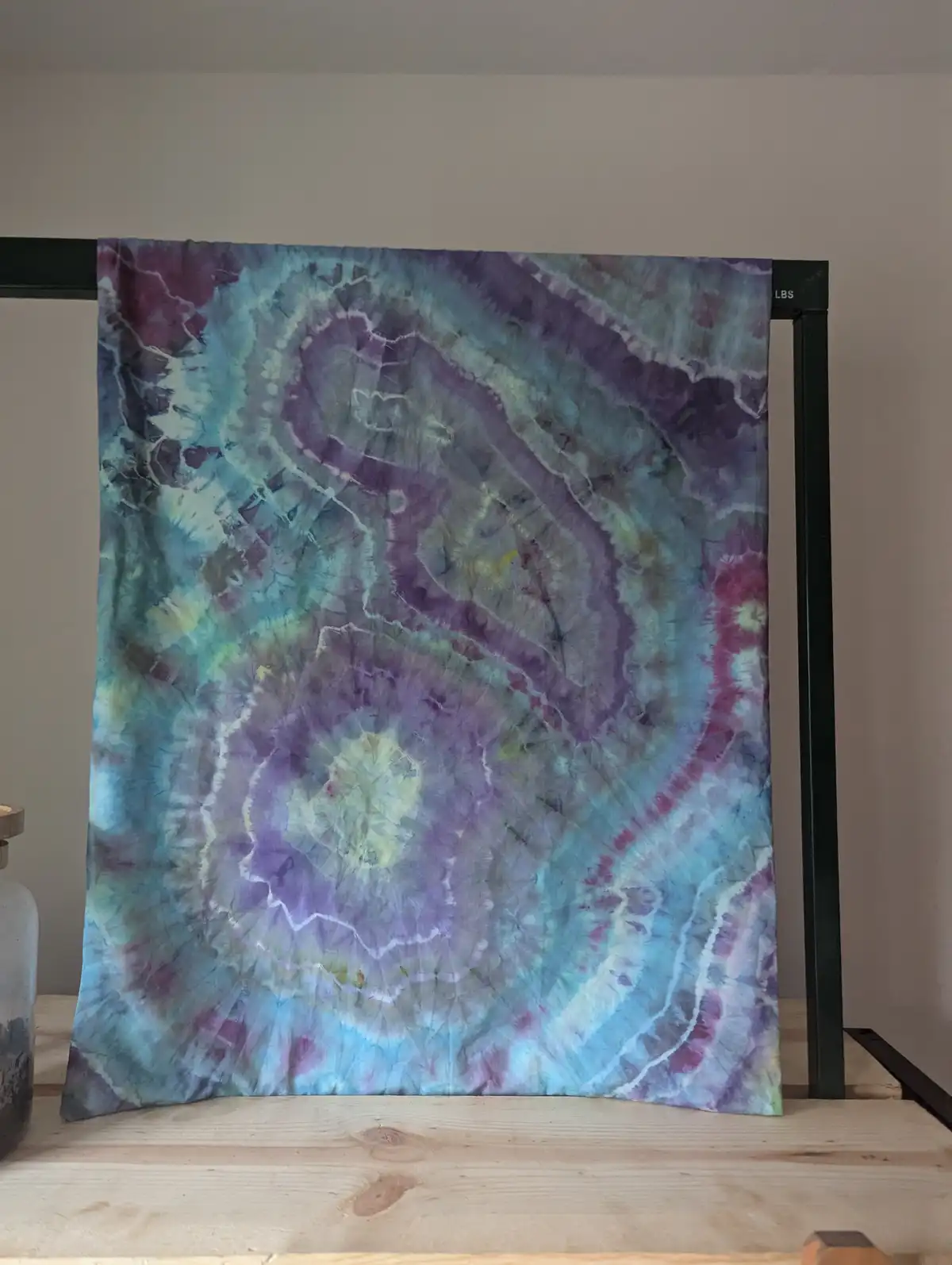 A vibrant tie-dye fabric with swirling patterns of purple, teal, and white hangs on a black metal rack against a plain wall…