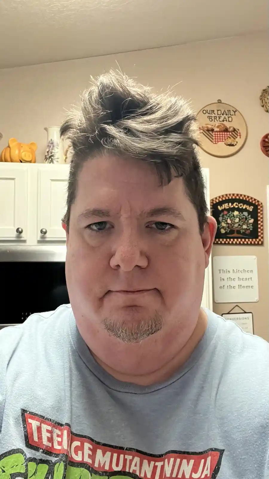 A man with messy, spiky hair and a stern, unamused facial expression stares directly at the camera. He wears a light blue…
