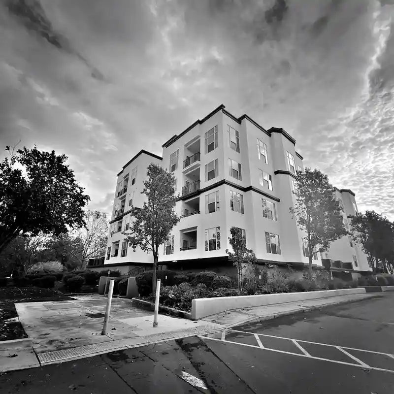 A black-and-white photo of a modern, multi-story apartment building under a cloudy sky, with trees and a sidewalk in the…