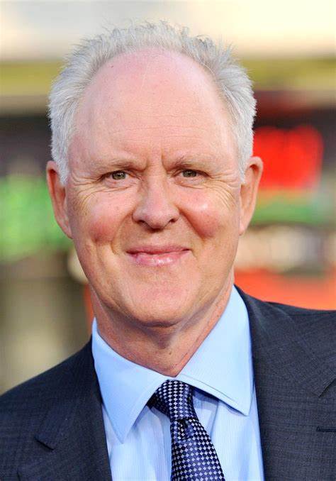A close-up photo of a smiling older white man with white hair, wearing a dark suit, light blue shirt, and patterned tie. The…