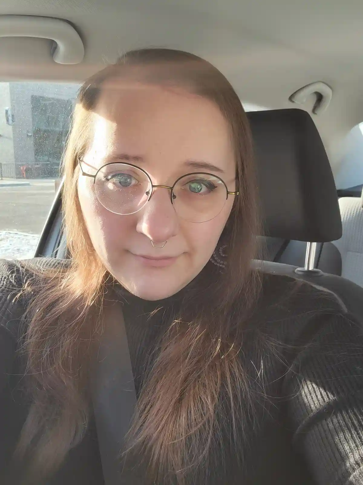 A person with glasses and a nose ring sits in a car, looking directly at the camera with a subtle, slightly smug expression…