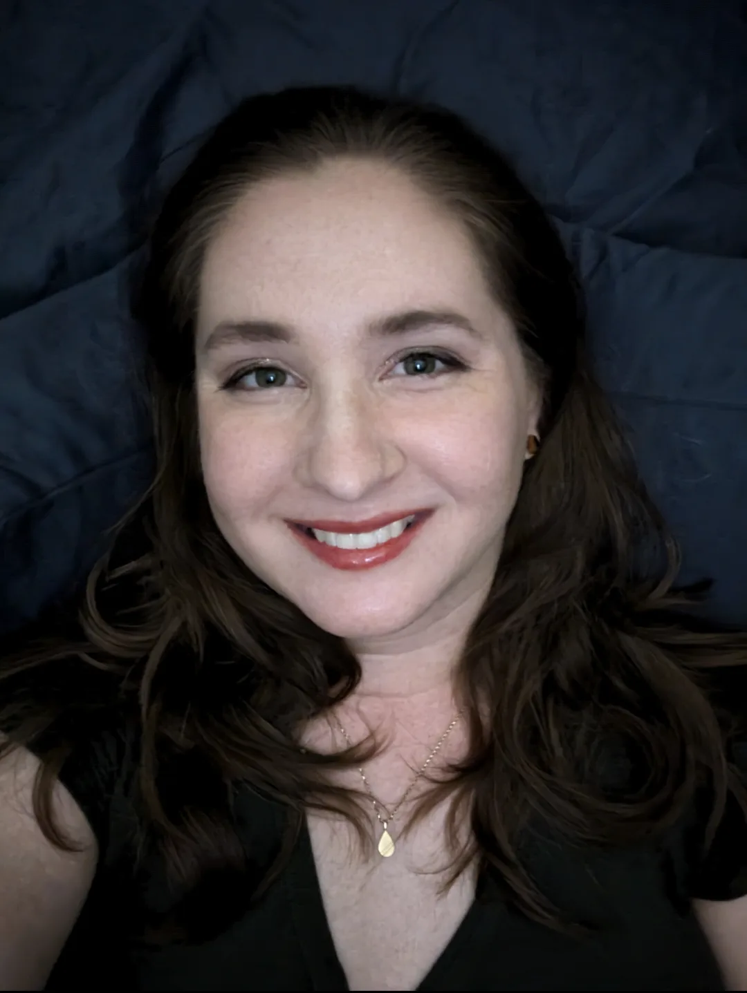 A woman with brown hair and a warm smile lies on a dark blue bedspread, wearing a black top and a gold teardrop pendant…