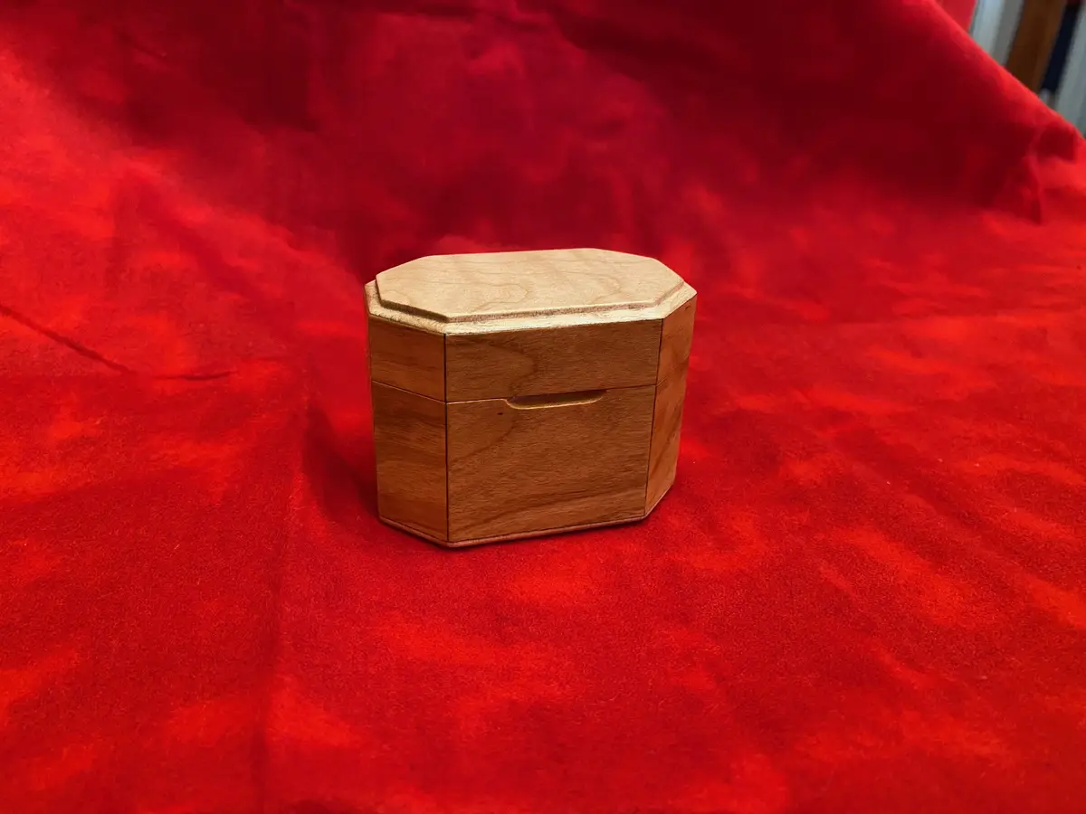 A small, octagonal wooden box with a smooth finish and a subtle groove on its lid sits centered on a rich, textured red…