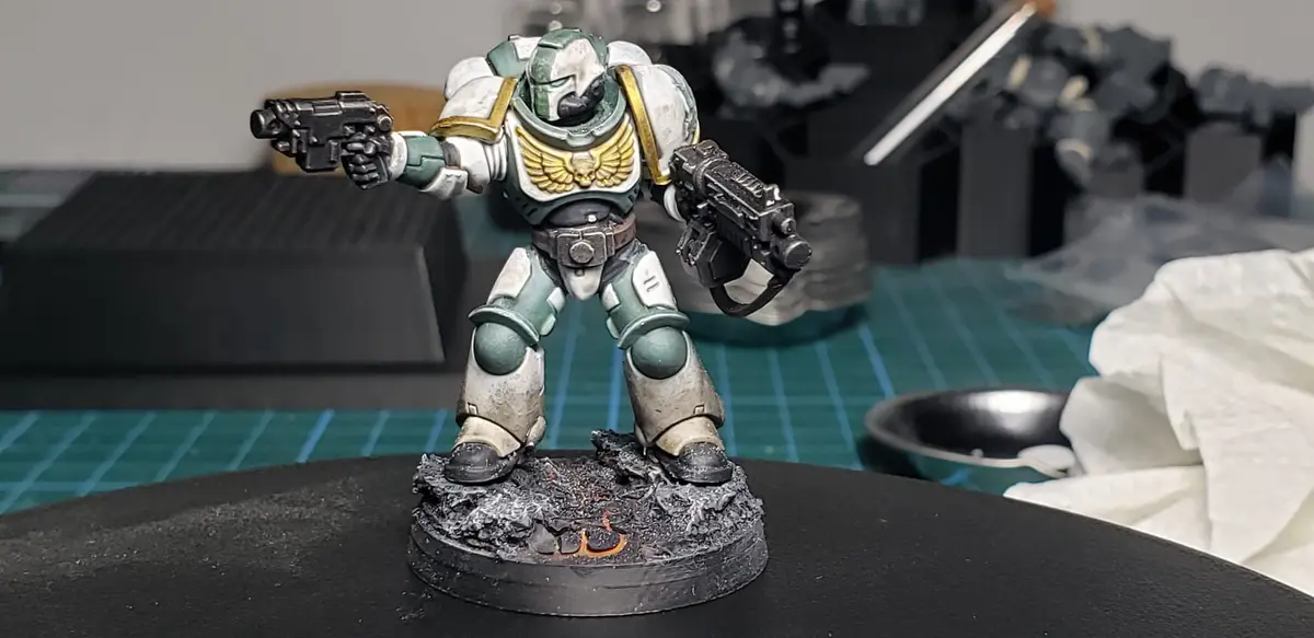 A painted miniature of a Space Marine in green and white armor with gold wings stands on a black base with orange flames…