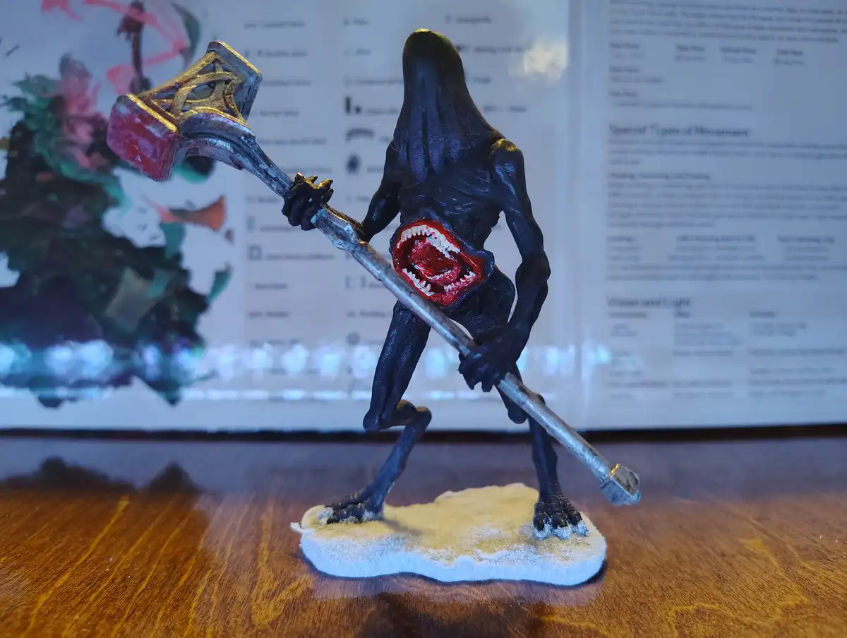 A painted miniature figurine of a dark, humanoid monster with a gaping, toothy maw holding a large ornate hammer stands on a…