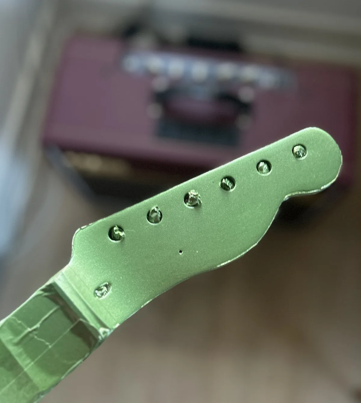 A close-up photo shows the headstock of a green electric guitar, with all six tuning peg holes empty and devoid of screws…