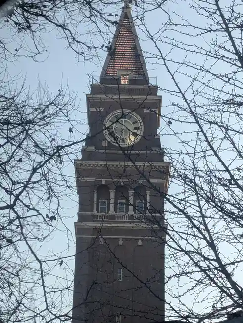 A tall brick clock tower with a large white-faced clock is viewed through the bare branches of trees against a pale sky. The…