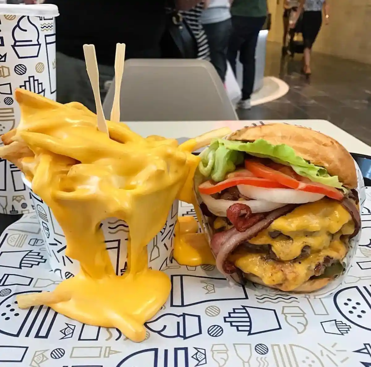 A close-up photo shows a large cheeseburger with lettuce, tomato, onion, bacon, and melted cheese, next to a cup of french…