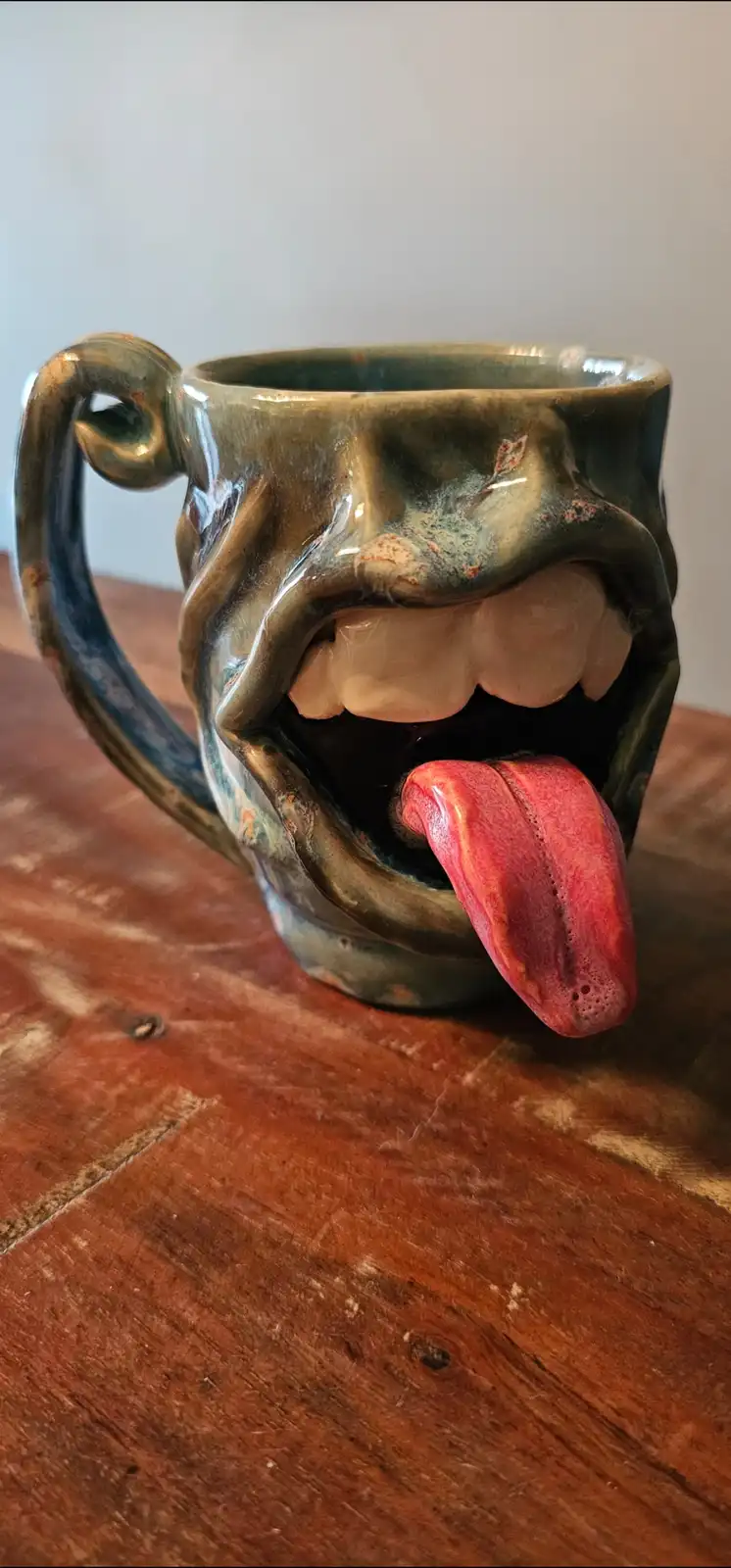 A ceramic mug shaped like a screaming face with an open mouth, protruding teeth, and a long red tongue sticking out, sits on…