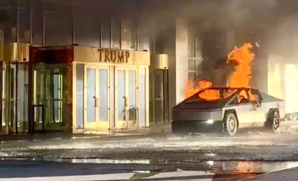 A white Tesla Cybertruck is engulfed in flames on a street at night, with thick smoke rising. Behind it, a building entrance…