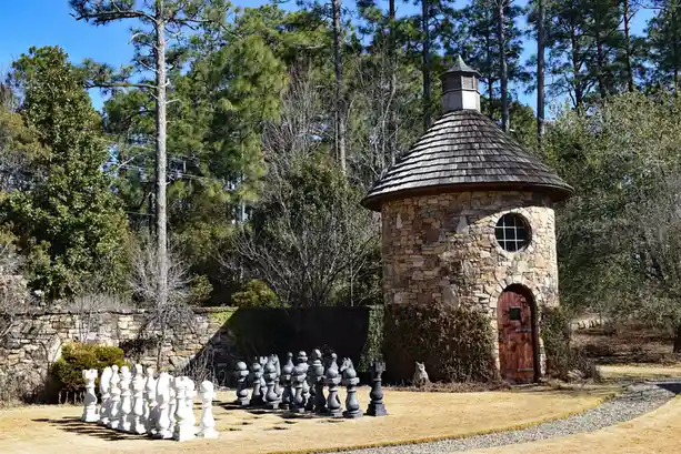 A large outdoor chess set with white and black pieces is arranged on dry grass in front of a stone gazebo with a wooden door…