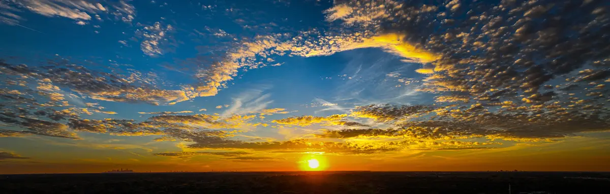 A panoramic photograph of a vibrant sunset with the sun low on the horizon, casting golden light on scattered clouds against…