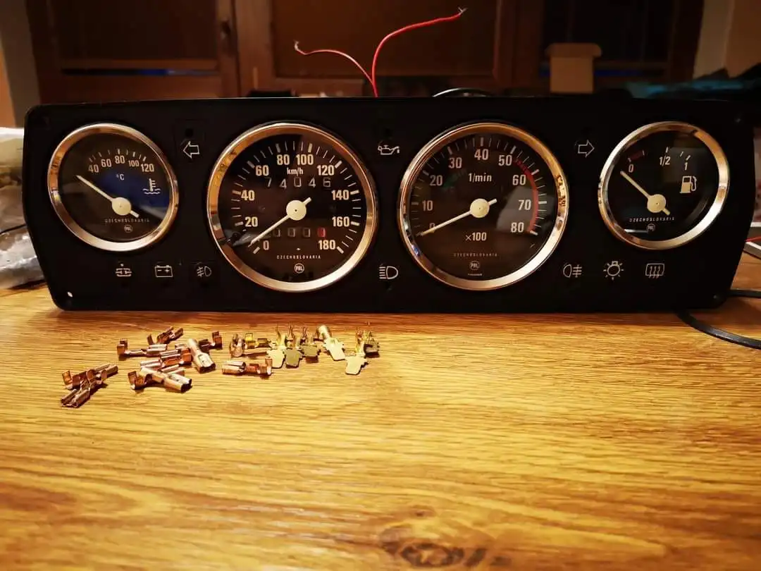 A vintage car dashboard with four analog gauges (temperature, speed, RPM, fuel) sits on a wooden table, with a pile of…