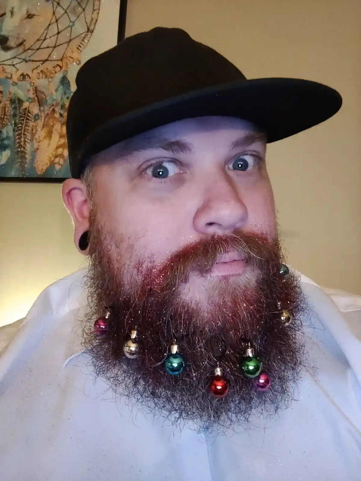 A man with a thick, reddish-brown beard adorned with colorful Christmas ornaments and glitter, wearing a black baseball cap…