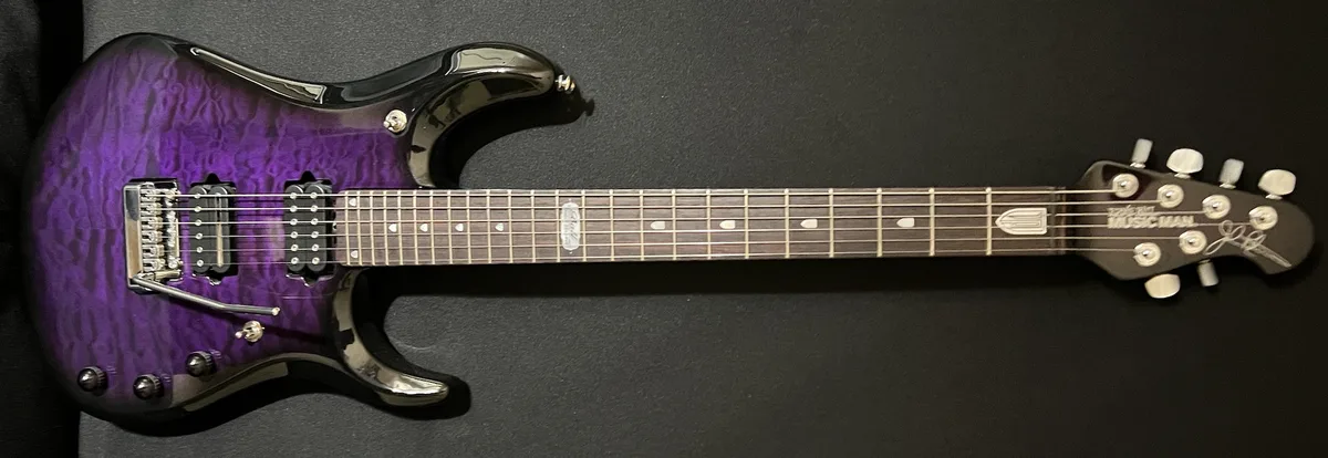 A purple and black electric guitar with a quilted finish lies horizontally on a dark surface. The headstock reads 'Music…
