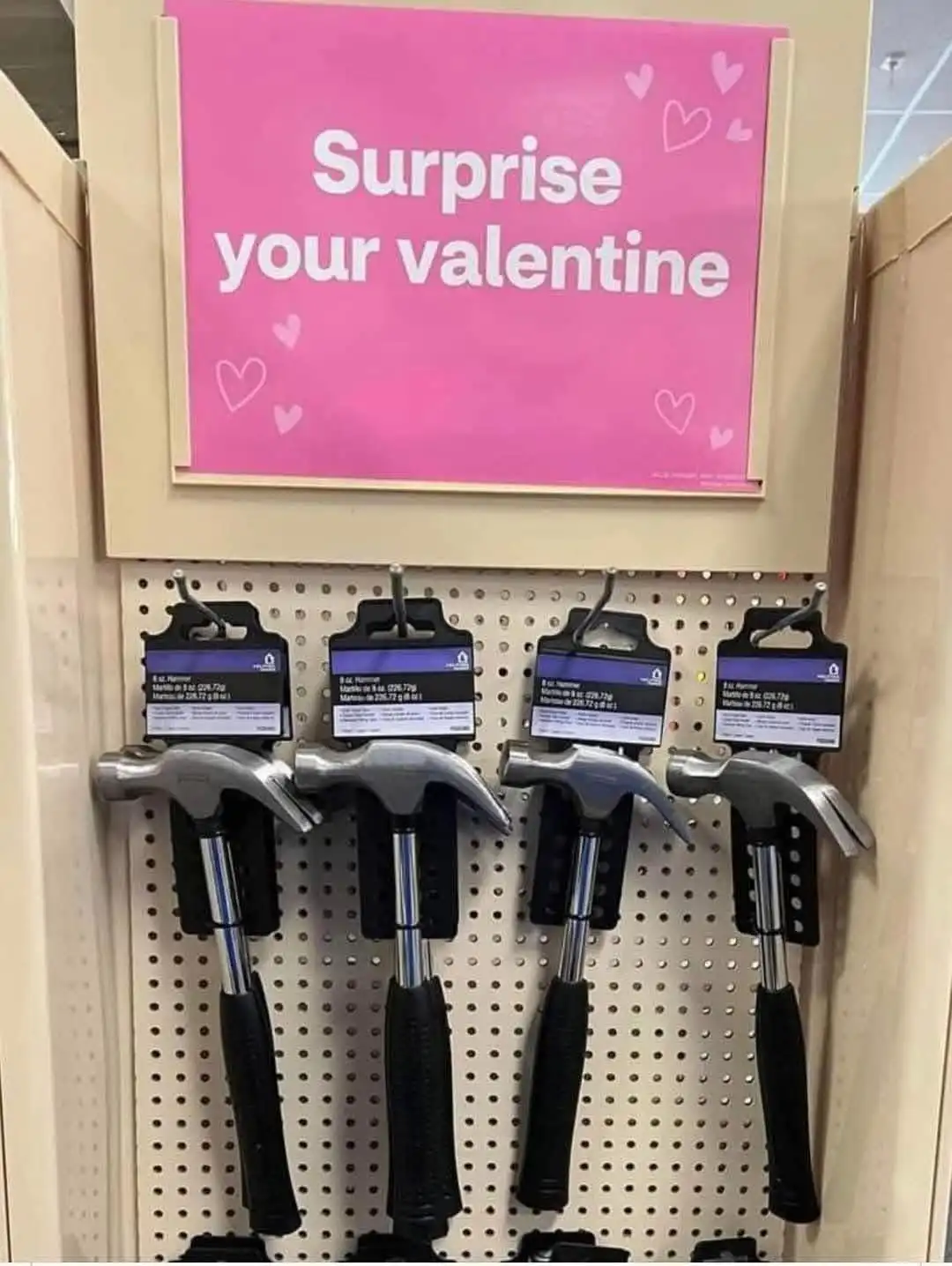 A retail display shows four identical 8-oz hammers hanging on a pegboard beneath a bright pink sign decorated with hearts…