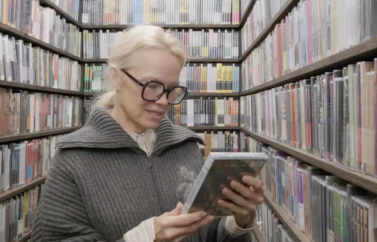 A woman with blonde hair and black-rimmed glasses stands in an aisle of a media store surrounded by shelves of CDs. She is…