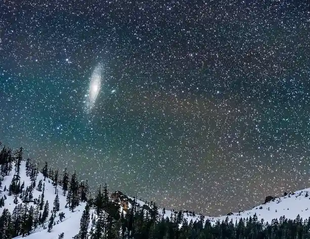 A stunning night sky photograph shows a snow-covered mountain range with dense pine forests in the foreground, under a vast…
