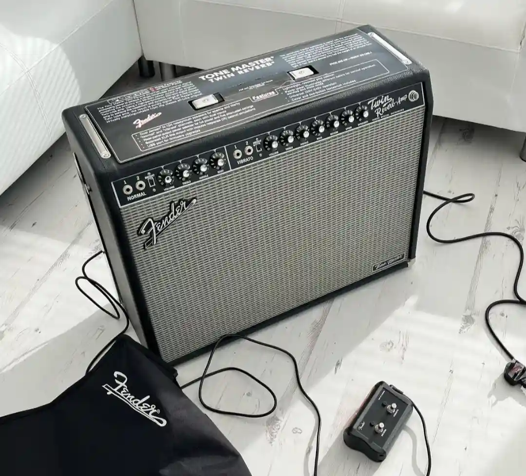 A black Fender Tone Master Twin Reverb guitar amplifier sits on a light-colored wooden floor next to a white couch, with a…