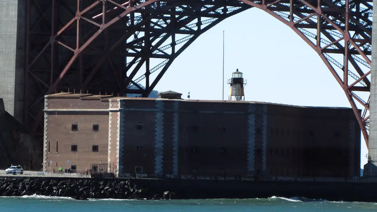 A photo shows a large brick fort under the red steel arches of the Golden Gate Bridge, with a small lighthouse on its roof…