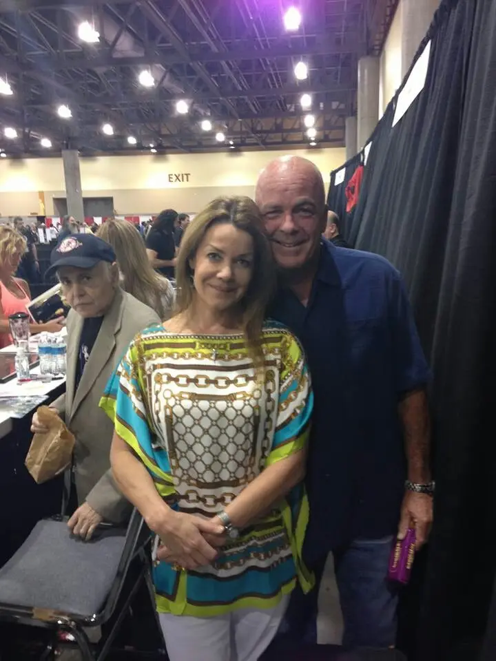 A photo taken at a convention or event shows a bald man in a blue shirt smiling broadly while standing next to a woman in a…