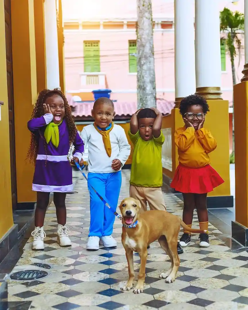 Four children in colorful outfits stand in a courtyard with a dog on a leash; the child holding the leash looks surprised…