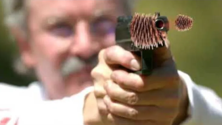 A man with a mustache holds a handgun pointed forward, but the barrel and a nearby pine cone are covered in spiky pine cone…