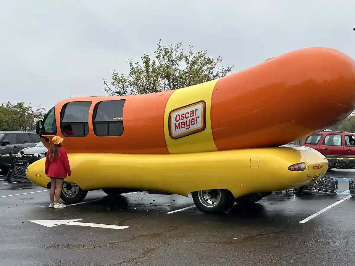 A person in a red jacket and yellow hat stands in a wet parking lot, looking up at the iconic Oscar Mayer Wienermobile…