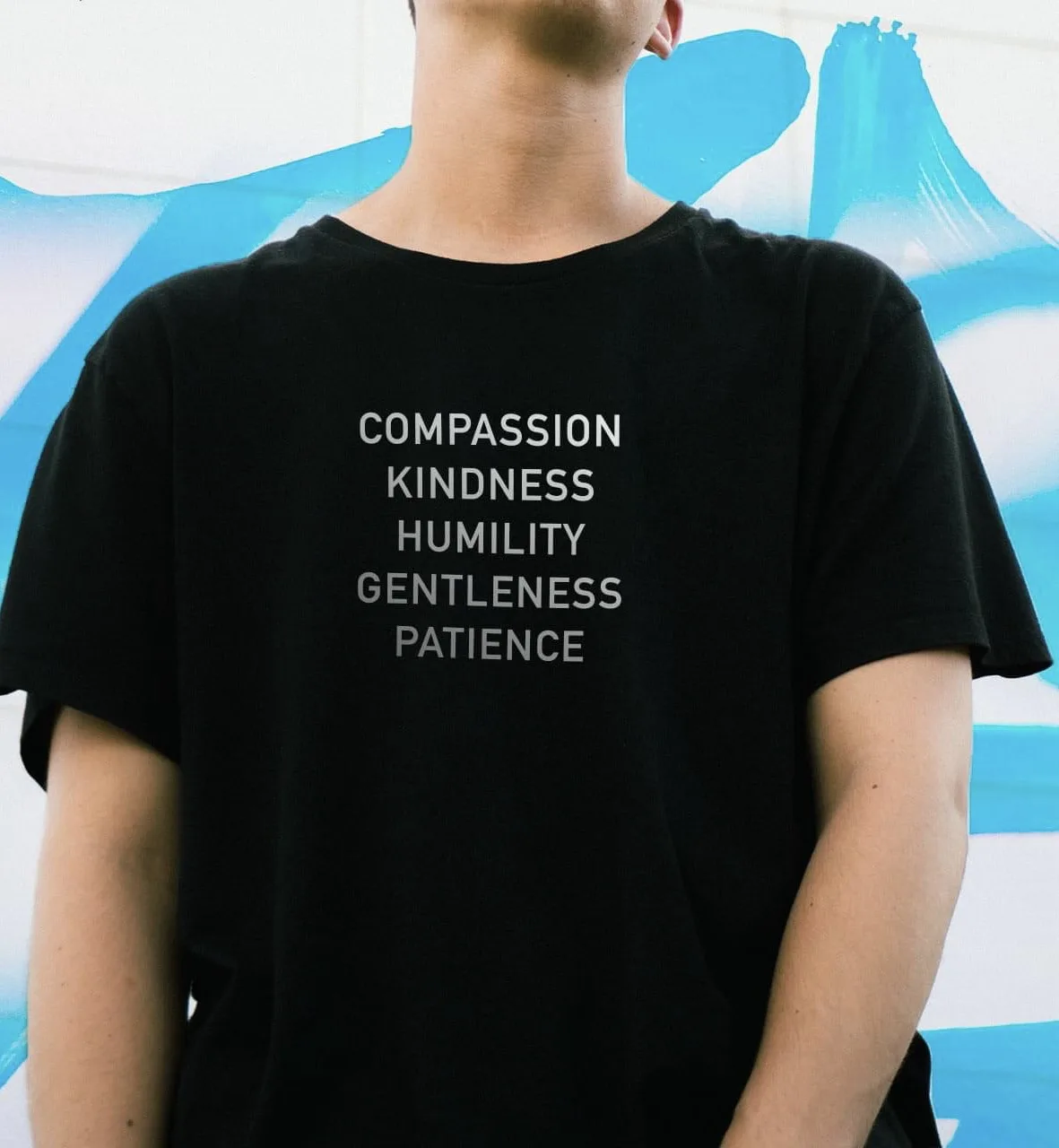 A person wearing a black t-shirt with five virtues—compassion, kindness, humility, gentleness, and patience—printed in white…