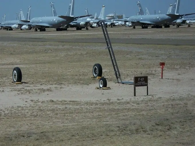 A desert airfield shows rows of parked military jets in the background. In the foreground, three airplane tires are propped…