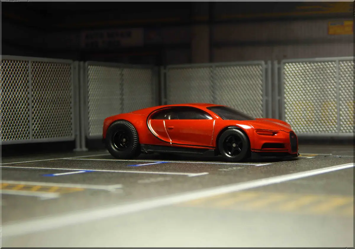 A small, red toy model of a Bugatti Chiron is parked in a miniature parking garage with white lines and chain-link fences in…