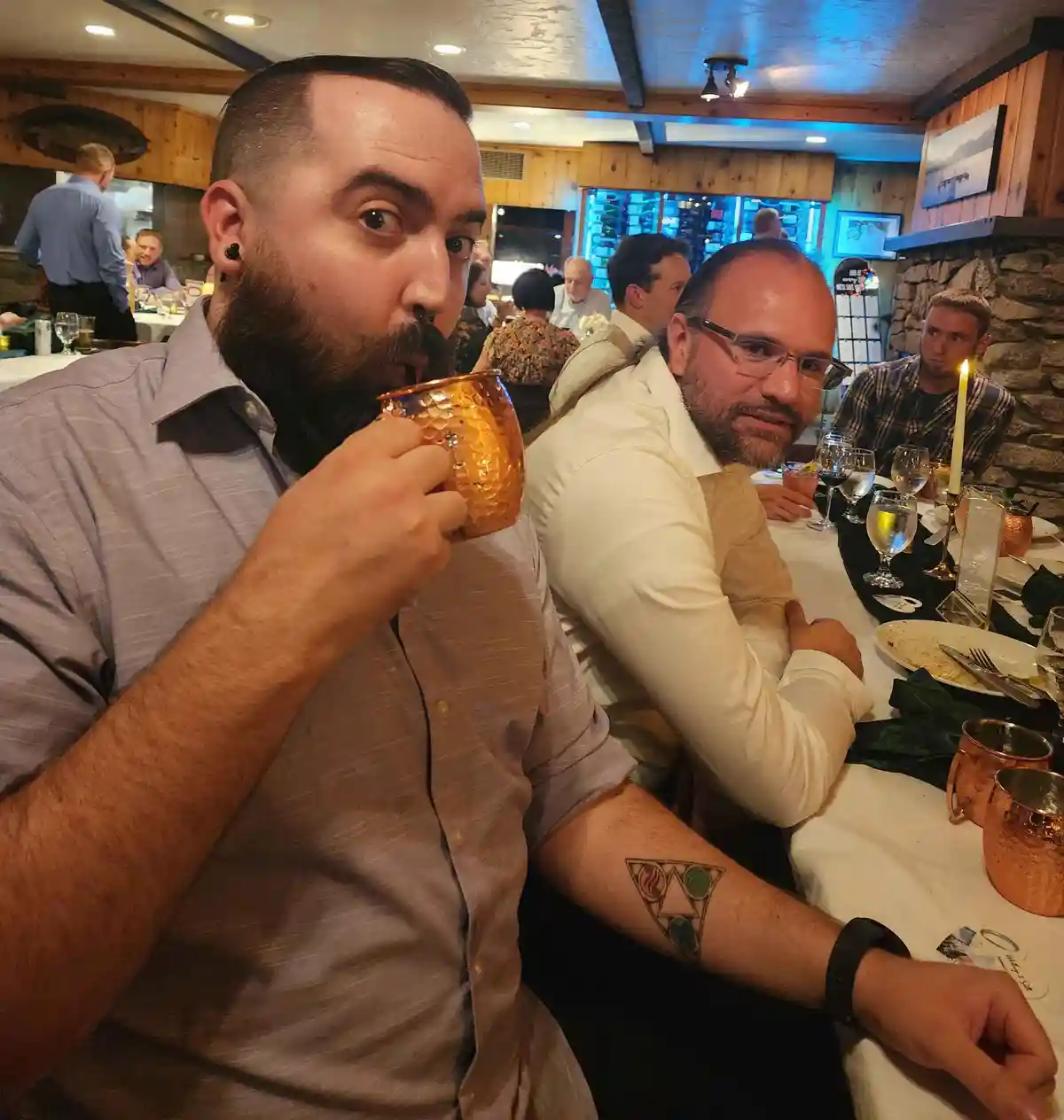 A man with a beard and nose ring drinks from a copper mug at a restaurant table, looking surprised. Beside him, another man…