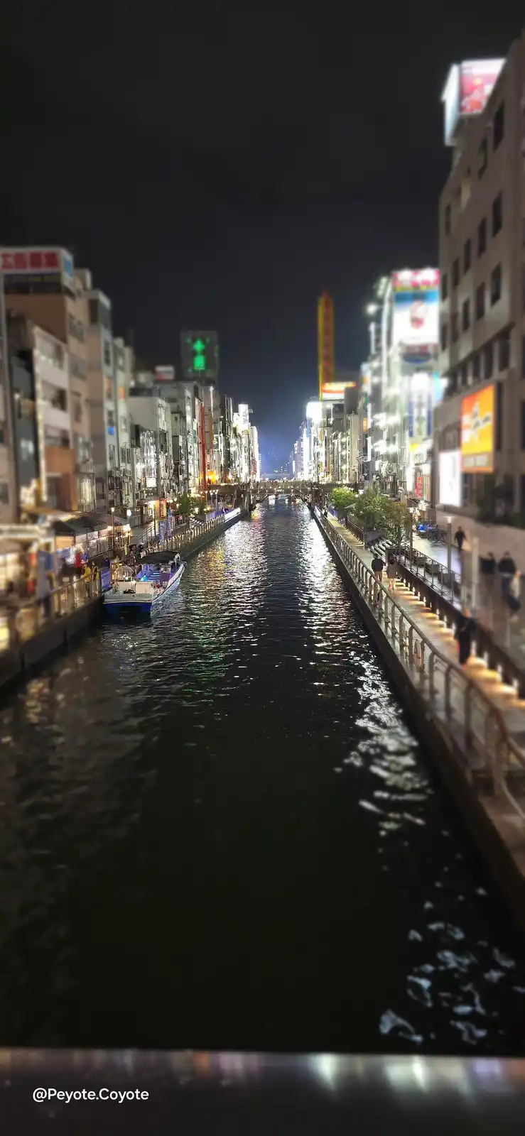 A nighttime photo of a narrow, illuminated canal in a dense urban area, flanked by tall buildings with bright neon signs and…