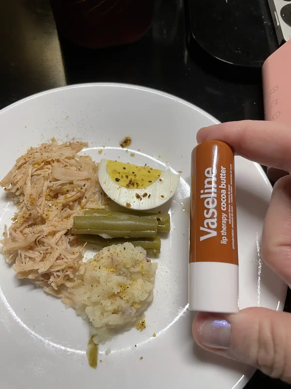 A hand holds a tube of Vaseline lip therapy cocoa butter next to a plate of food, which includes mashed potatoes, green…