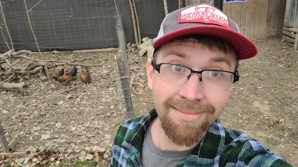 A man with glasses and a plaid shirt takes a selfie in front of a wire fence, behind which several chickens roam in a yard…