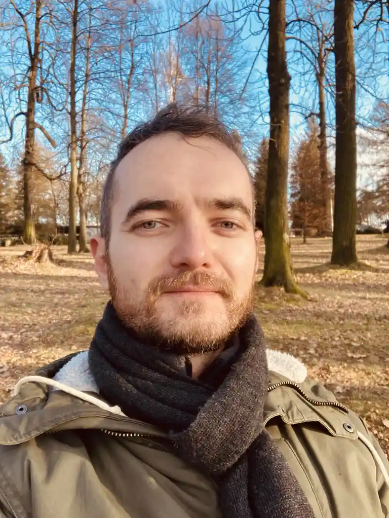 A man with a beard and short hair takes a selfie outdoors in a park with bare trees and fallen leaves under a clear blue…