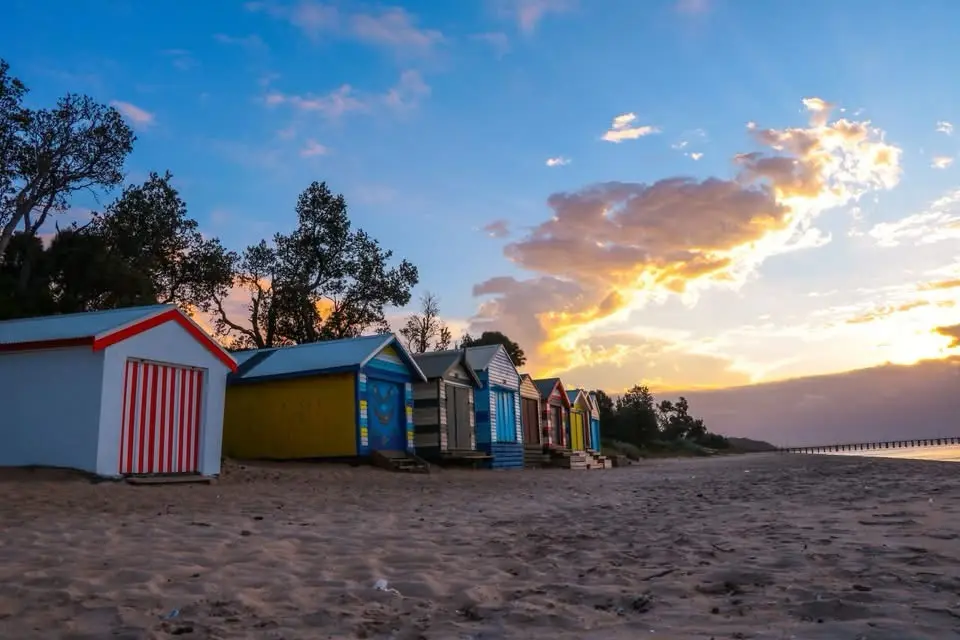 A row of colorful beach huts on a sandy shore at sunset, with trees in the background and a pier visible in the distance…