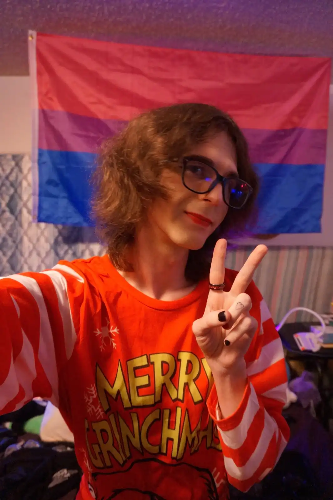 A person with curly brown hair and glasses, wearing a red and white striped 'Merry Grinchmas' shirt, makes a peace sign with…