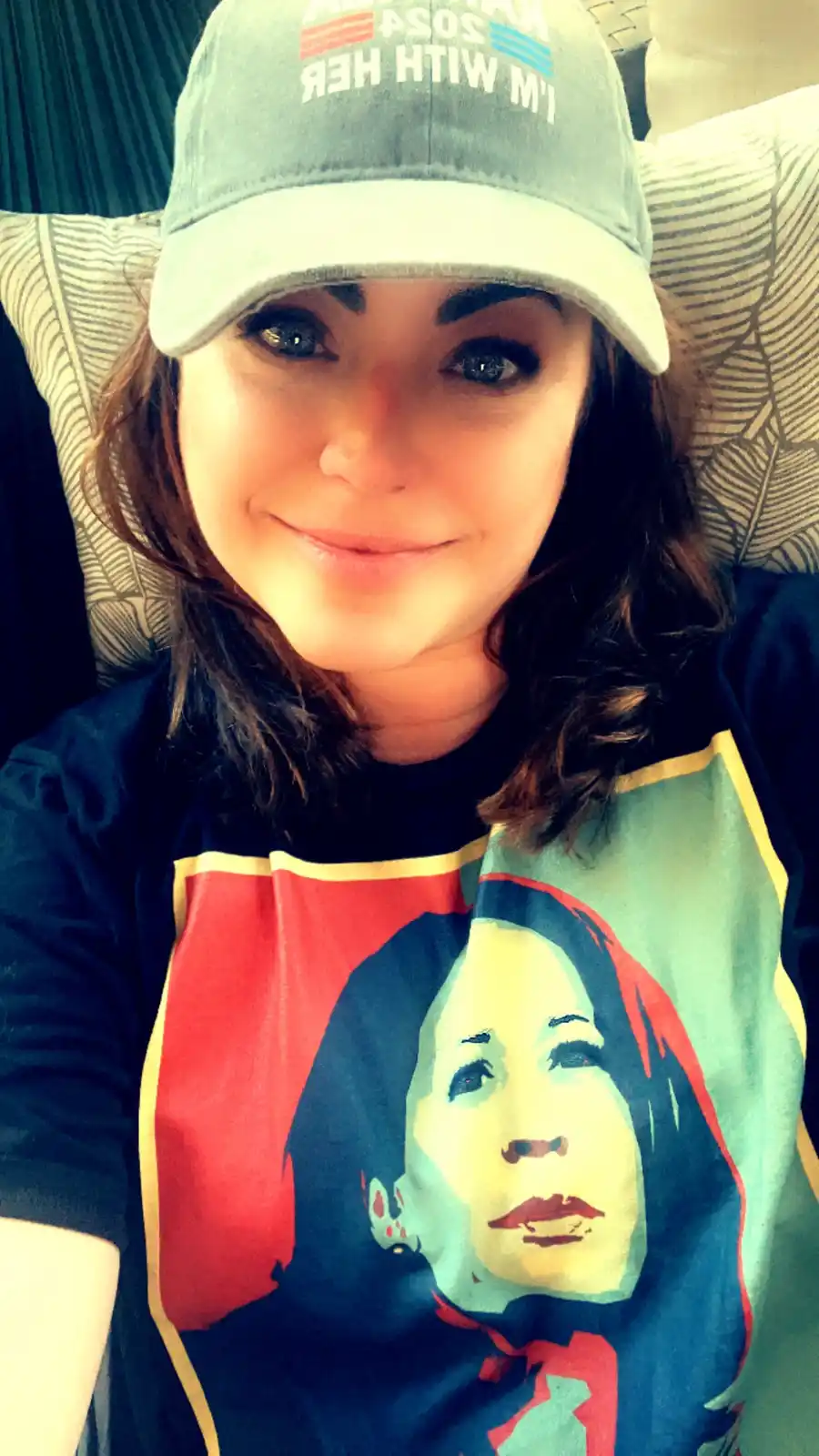 A woman with dark hair and a slight smile wears a light blue baseball cap that reads 'I'M WITH HER 2024' and a dark t-shirt…