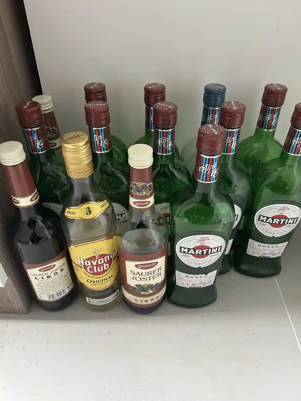 A collection of liquor bottles, including multiple green Martini Rosso bottles, a Havana Club rum, and two Saurer Joster…