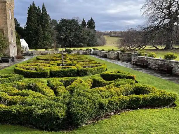 A photograph of a formal garden with neatly trimmed hedges arranged in a complex, winding maze pattern, surrounded by stone…
