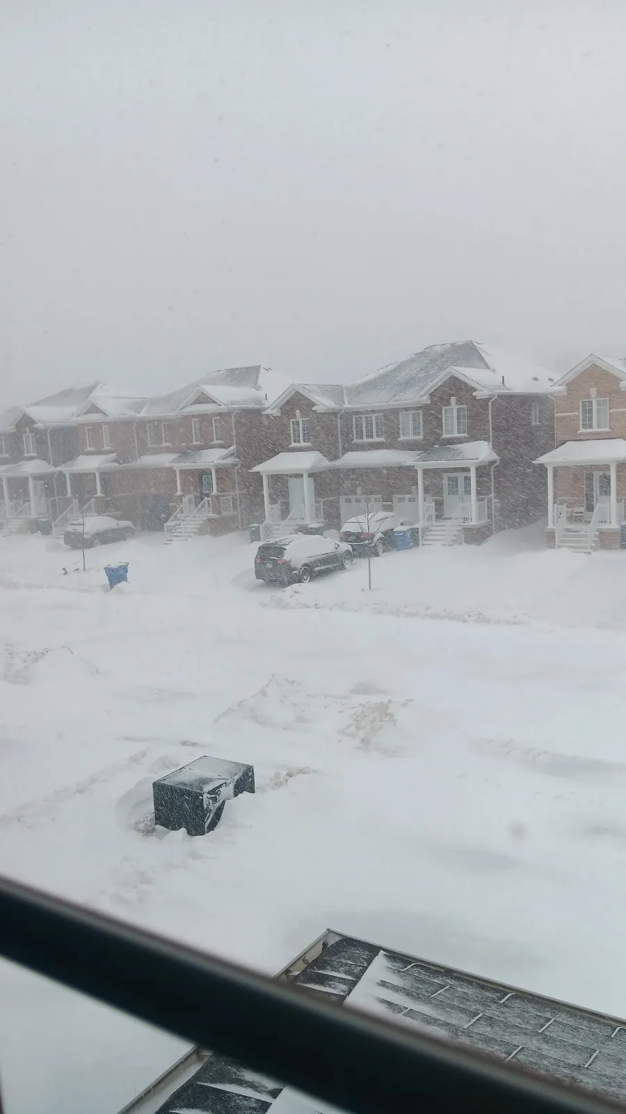 A heavy snowstorm blankets a residential neighborhood, with snow-covered brick townhouses, parked cars, and a black outdoor…