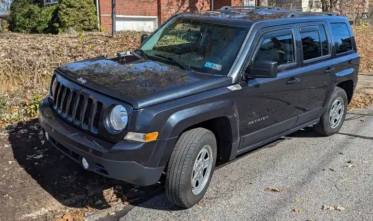 A dark gray Jeep Patriot SUV is parked on a gravel driveway next to a leaf-covered bush, with a brick house in the…
