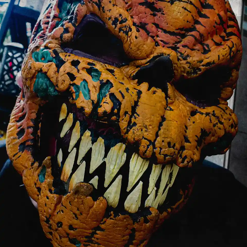 A close-up of a person wearing a grotesque, orange and black monster mask with exaggerated, jagged teeth. The mask has a…